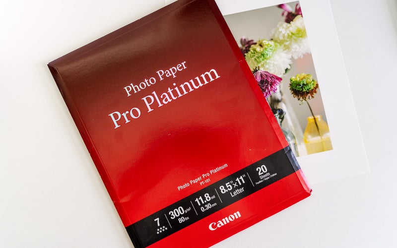 A red package of Canon Pro Platinum photo paper with a printed photograph of flowers underneath rests on a white desk.